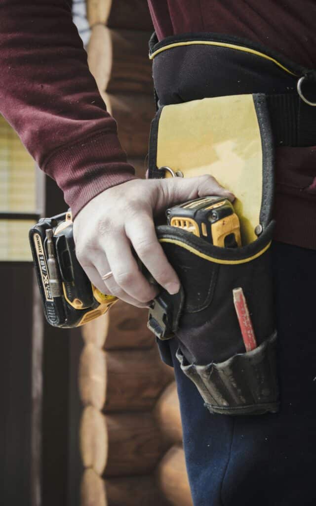 Person wearing a tool belt holding a power drill, emphasizing tools for smoke and carbon monoxide detector maintenance and battery replacement.