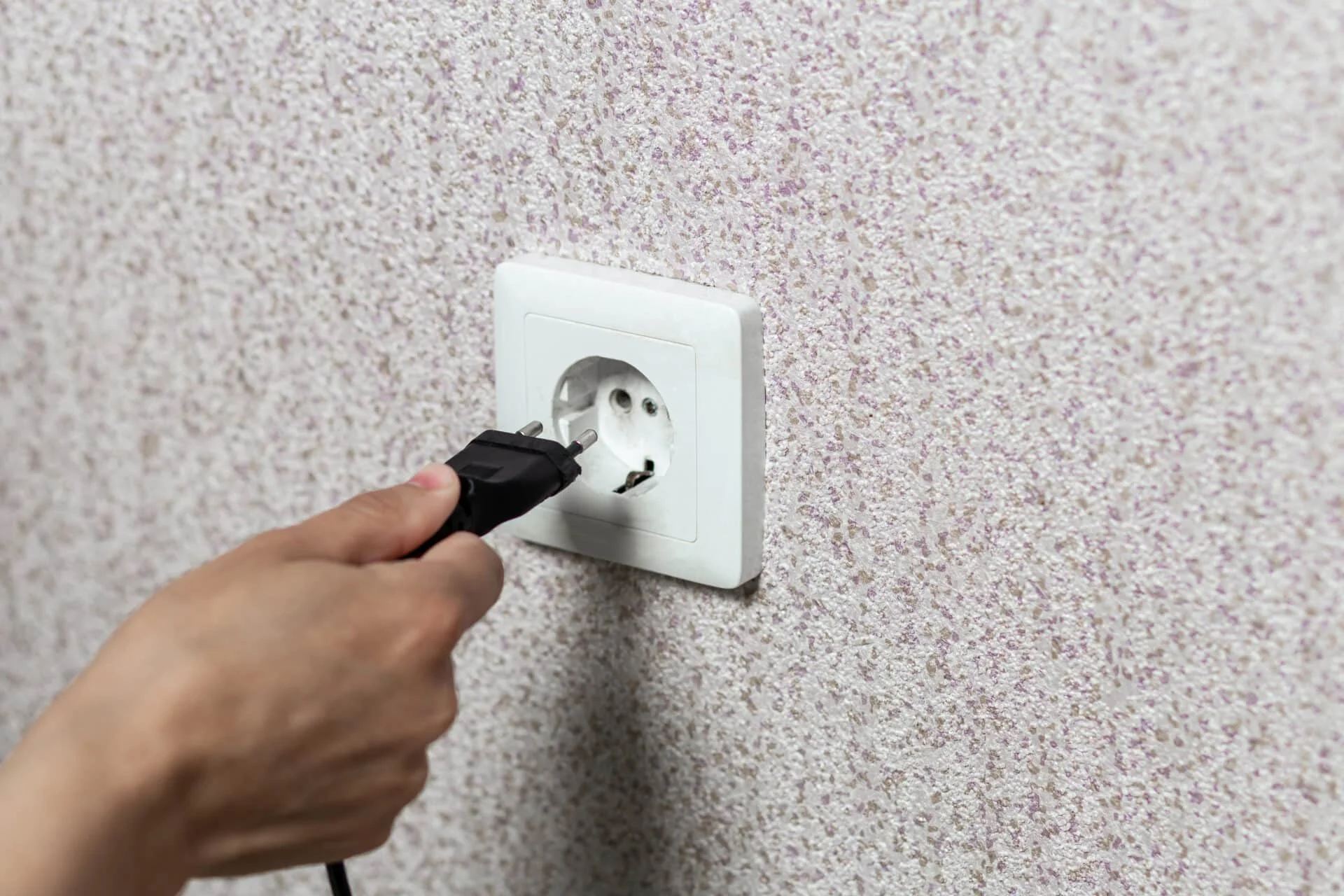 Hand plugging a black electrical cord into a white outlet on textured wall, illustrating proper outlet installation for electrical safety and functionality in homes.