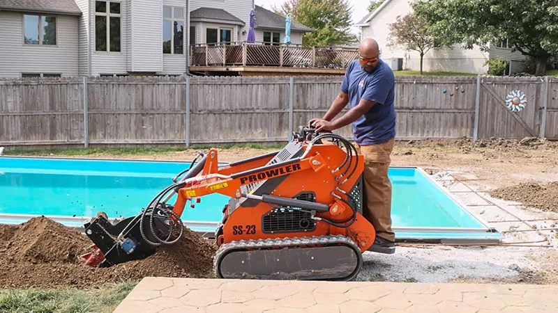 Man operating a Prowler SS-23 mini excavator near a pool, digging and preparing the ground for outdoor lighting installation in a residential backyard.